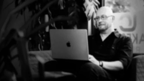 Ein Mann sitzt auf einem Sofa und arbeitet an einem Laptop. Er trägt eine schwarze Kleidung und schaut konzentriert auf den Bildschirm. Im Hintergrund sind Pflanzen und ein Fenster zu sehen, das Tageslicht hereinlässt.