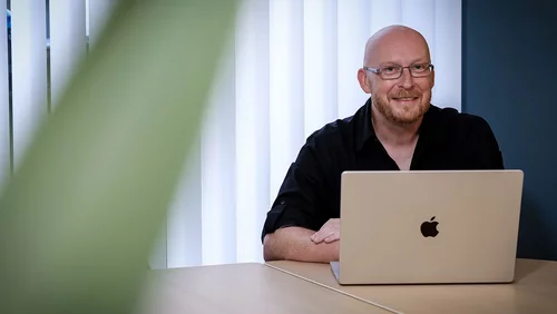 Ein lächelnder Mann sitzt an einem Tisch vor einem Laptop, auf dem ein Apfel-Logo sichtbar ist. Er trägt ein schwarzes Hemd und Brille. Im Hintergrund sind helle Fenster mit Jalousien zu sehen.