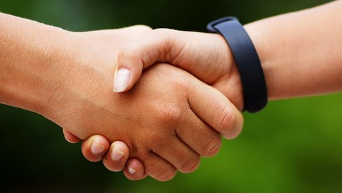 Zwei Hände schütteln sich fest, symbolisieren Zusammenarbeit und Freundschaft. Eine Hand hat einen schwarzen Fitness-Tracker am Handgelenk. Der Hintergrund zeigt unscharfe grüne Elemente, die eine natürliche Umgebung andeuten.