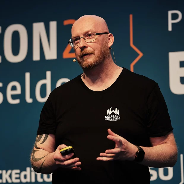 Wolfgang Wagner steht auf einer Bühne bei der T3CON in Düsseldorf. Er trägt ein schwarzes T-Shirt und hält ein Gelb gefärbtes Präsentationswerkzeug in der Hand, während er spricht. Im Hintergrund ist das Event-Logo und einige Sponsoren sichtbar.