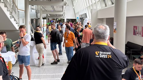 Teilnehmer und Aussteller bei den TYPO3 Developer Days 2024 in Karlsruhe versammeln sich in einem offenen Bereich mit verschiedenen Ständen und Roll-ups.