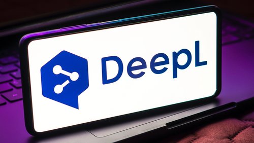 Auf dem Bild ist ein Smartphone zu sehen, das das Logo von DeepL zeigt. Das Logo besteht aus einem stilisierten Sprechblasen-Symbol und dem Schriftzug "DeepL" in dunkler Schrift auf hellem Hintergrund. Das Smartphone steht auf einem Laptop-Tastatur, die teilweise sichtbar ist.