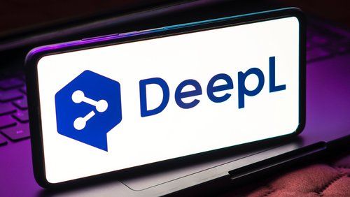Auf dem Bild ist ein Smartphone zu sehen, das das Logo von DeepL zeigt. Das Logo besteht aus einem stilisierten Sprechblasen-Symbol und dem Schriftzug "DeepL" in dunkler Schrift auf hellem Hintergrund. Das Smartphone steht auf einem Laptop-Tastatur, die teilweise sichtbar ist.