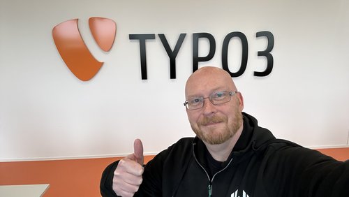 Wolfgang Wagner zeigt Daumen hoch vor einer Wand mit dem Schriftzug „TYPO3“ in Schwarz und dem orangefarbenen TYPO3 Logo. Der Hintergrund ist hell und freundlich.