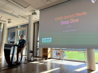 Ein Mann steht an einem Rednerpult und präsentiert. Im Hintergrund ist eine Leinwand mit dem Titel „TYPO3 Content Blocks Deep Dive“ und dem Namen „André Kraus“ zu sehen. Die Umgebung wirkt hell und modern, mit großen Fenstern.