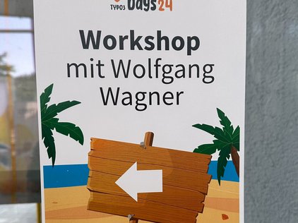 Türschild bei den TYPO3 Developer Days 2024 in Karlsruhe: Workshop mit Wolfgang Wagner. Der Pfeil zeigt nach links, das Schild ist mit Palmen und einem Strandmotiv dekoriert.